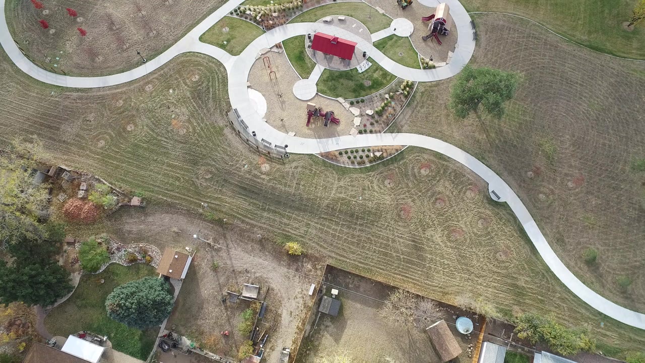 Overhead drone-style view in Hidden Lake, Colorado showing pale curved paths around mowed grass, a red-roofed park structure, playground equipment, nearby yards, and no visible people.