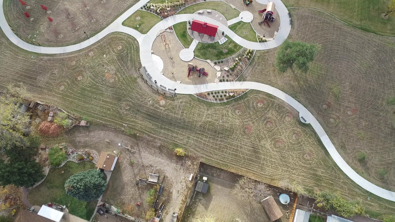 High overhead view in Hidden Lake, Colorado with winding walkways crossing open grass, a red-roofed shelter near playground features, yard edges, trees, and no visible people.