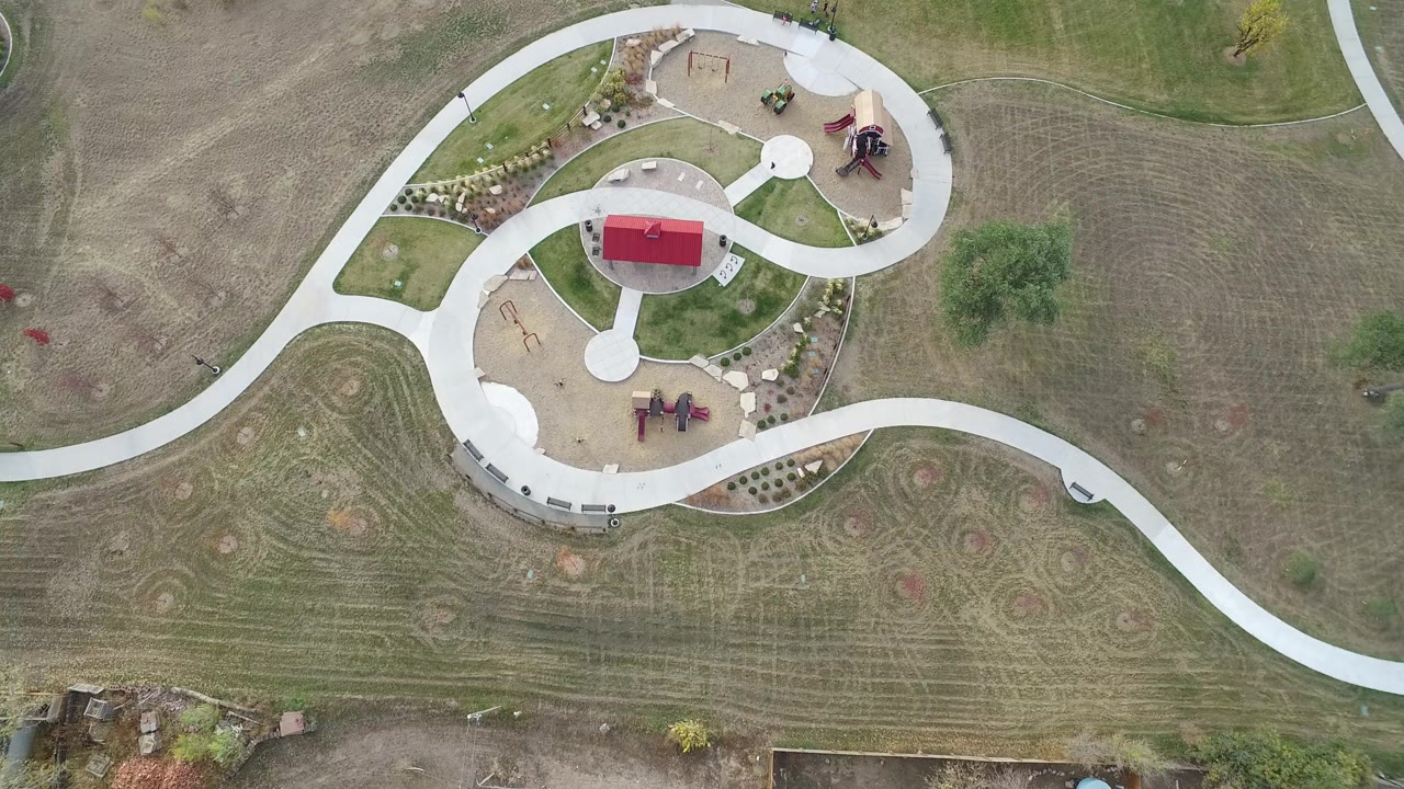 Overhead frame in Hidden Lake, Colorado showing pale curving walkways around a red-roofed shelter, tan play areas, planted beds, mowed open ground, and no visible people.