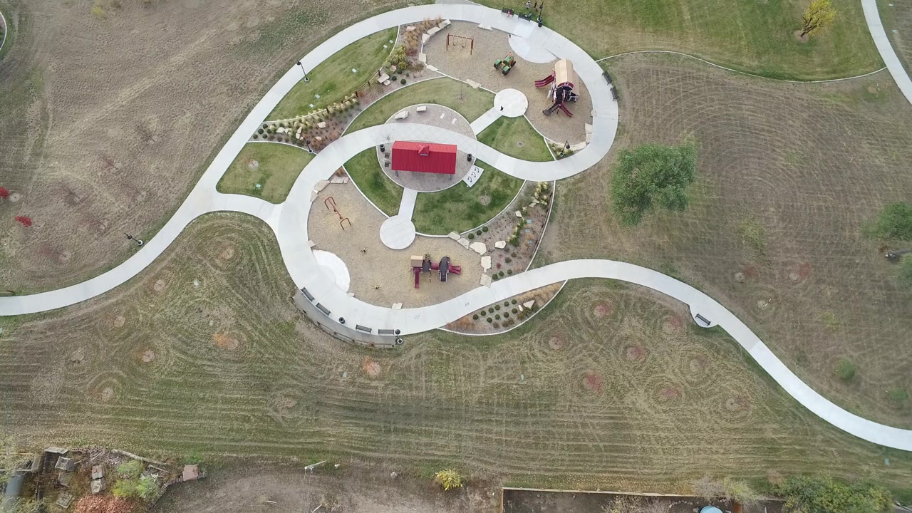 Hidden Lake, Colorado aerial frame with the red-roofed shelter centered among looping paths, tan playground surfaces, landscaped planting strips, and no visible people.