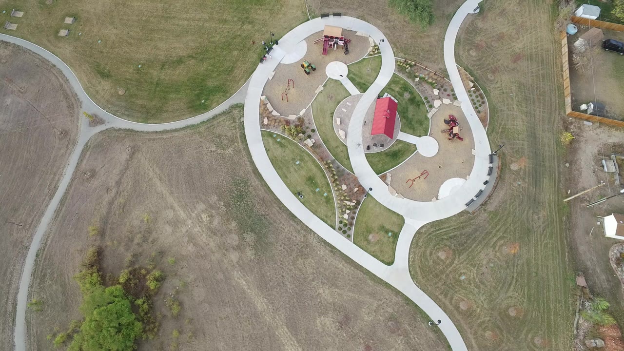 Overhead frame from Hidden Lake, Colorado showing pale curving sidewalks, a small playground area with red-roofed structures, open mowed land, scattered trees, and no visible people.