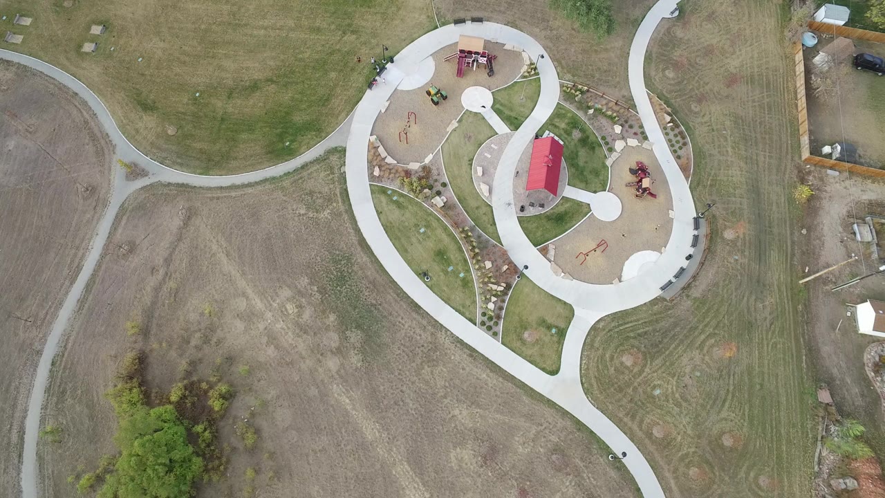 Second overhead frame in Hidden Lake, Colorado with the red-roofed park shelter near the upper right, looping concrete paths, tan play areas, mowed grass, and no visible people.