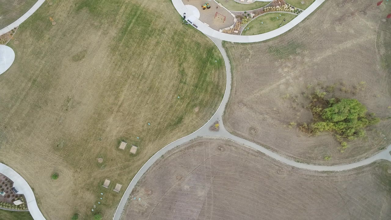 Overhead aerial frame from Hidden Lake, Colorado showing green and brown grass, pale curving paths, a small tree cluster near the right side, and no visible people.