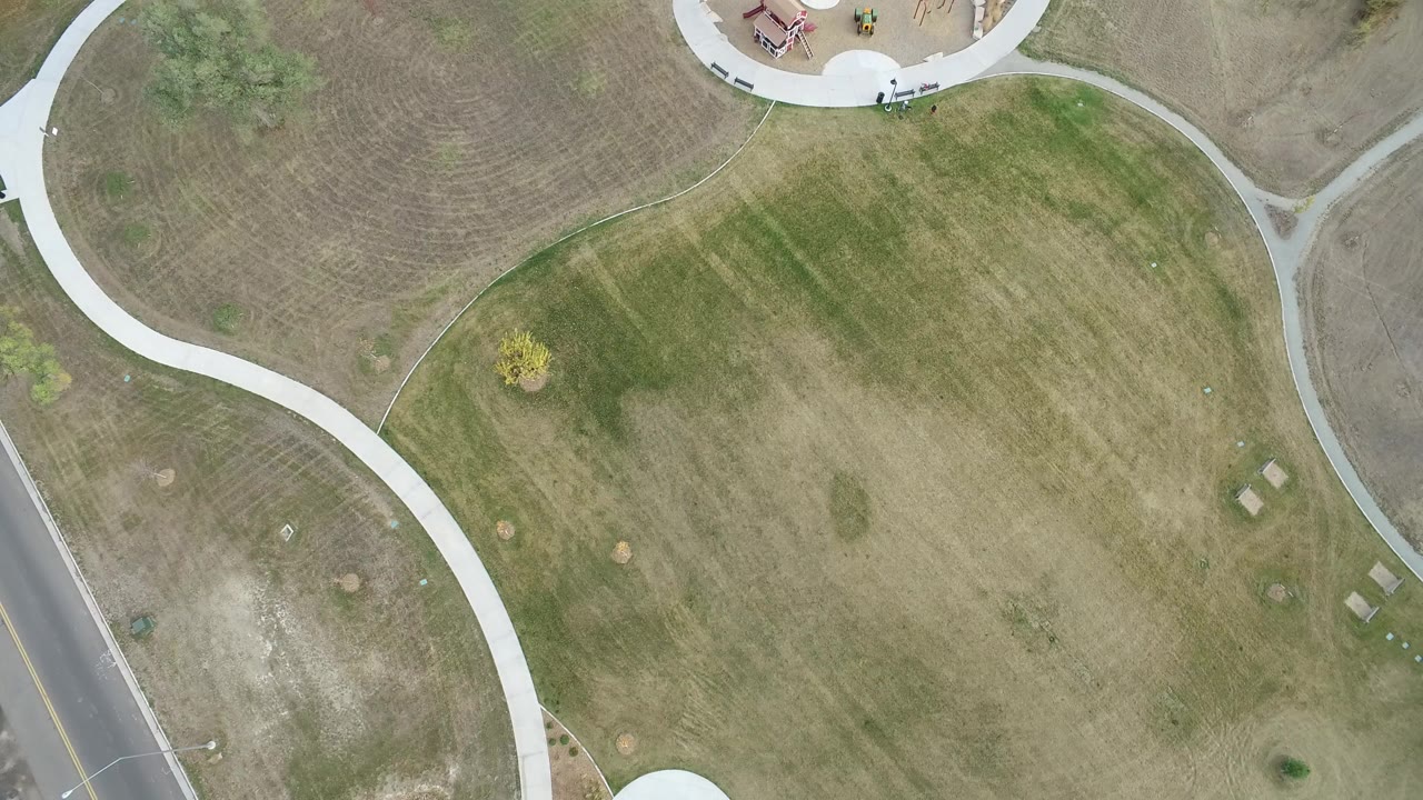 Top-down view of a Hidden Lake, Colorado park with curving pale walkways, patchy green-brown grass, a road at the left edge, and no visible people.
