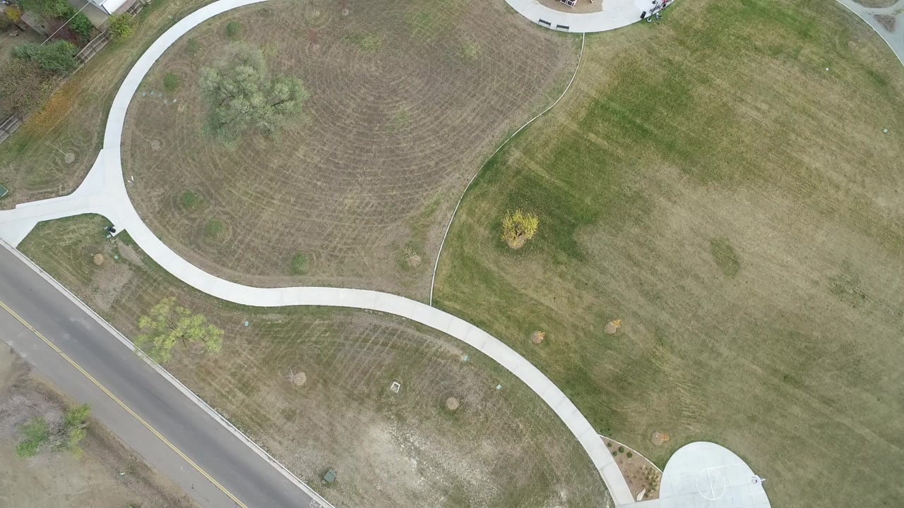 Overhead drone frame in Hidden Lake, Colorado showing pale curved paths across patchy grass, a paved road at lower left, scattered shrubs, and no visible people.