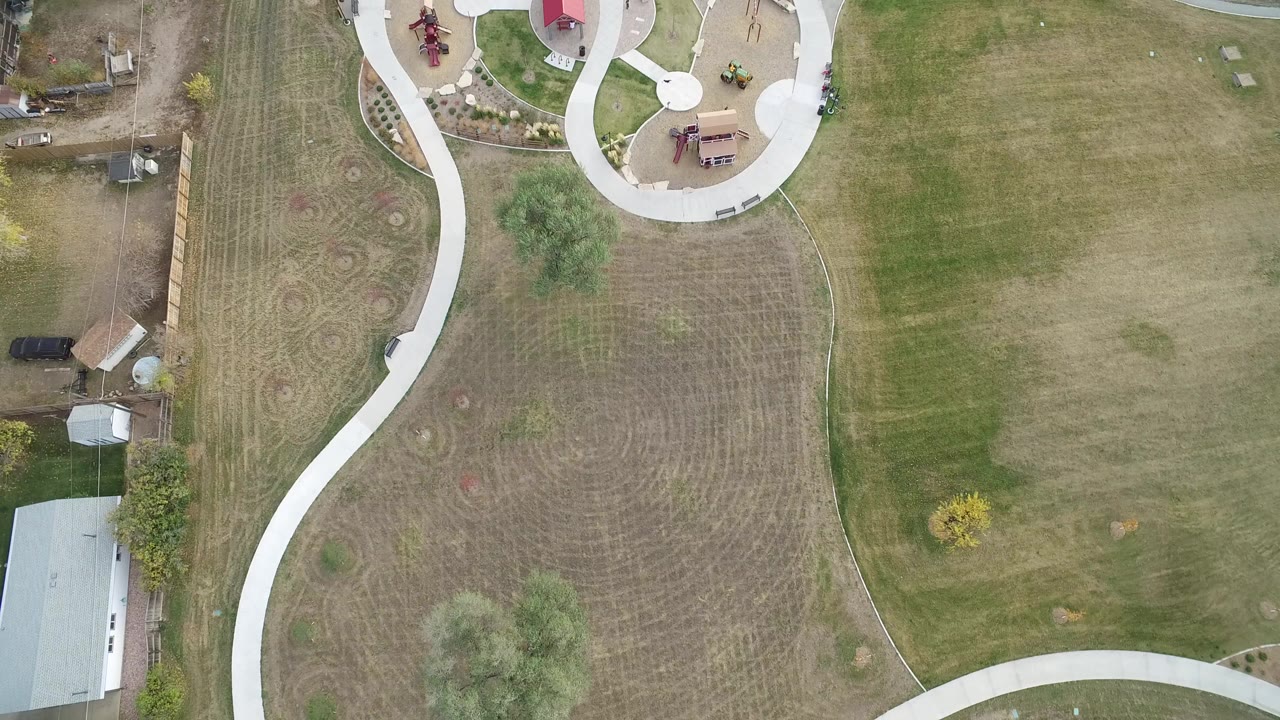 Top-down aerial frame in Hidden Lake, Colorado showing curving white sidewalks, mowed grass, garden beds, a red-roofed playground area, nearby outbuildings, and no visible people.