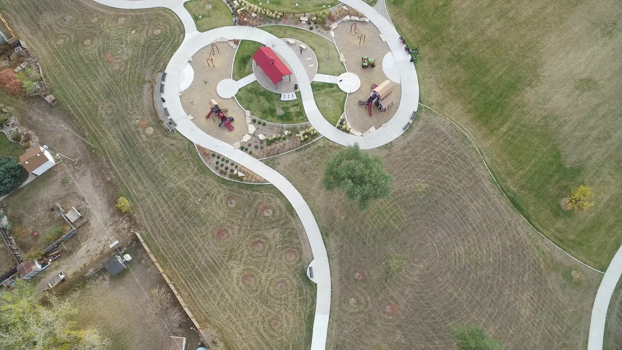 High overhead view in Hidden Lake, Colorado with the circular path network and red-roofed park structures near the upper frame and graded construction parcels along the lower-left side, with no visible people.