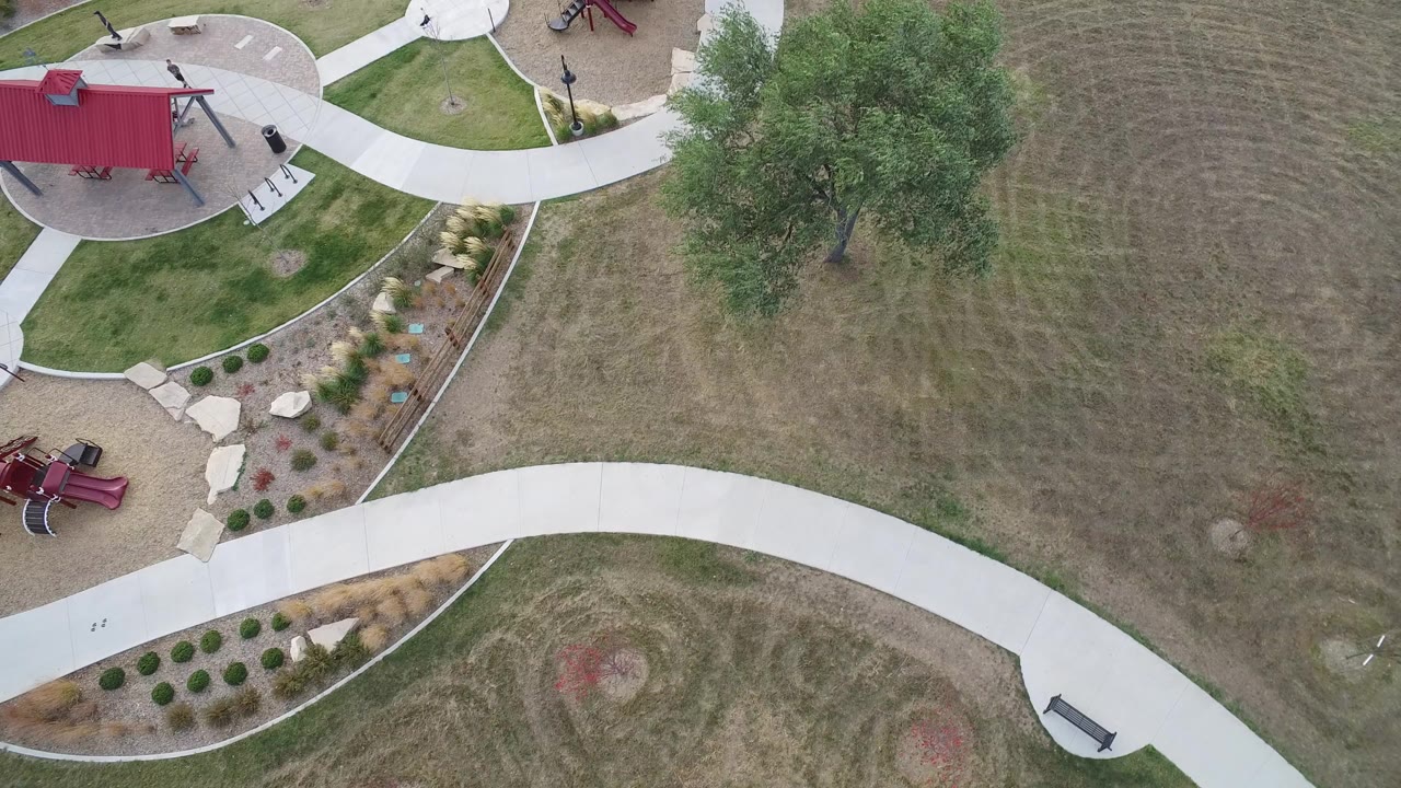 High overhead frame in Hidden Lake, Colorado showing curved concrete paths, brown-green mowed grass, a red park structure, playground equipment at the edges, a large tree, and tiny people visible on the grounds.