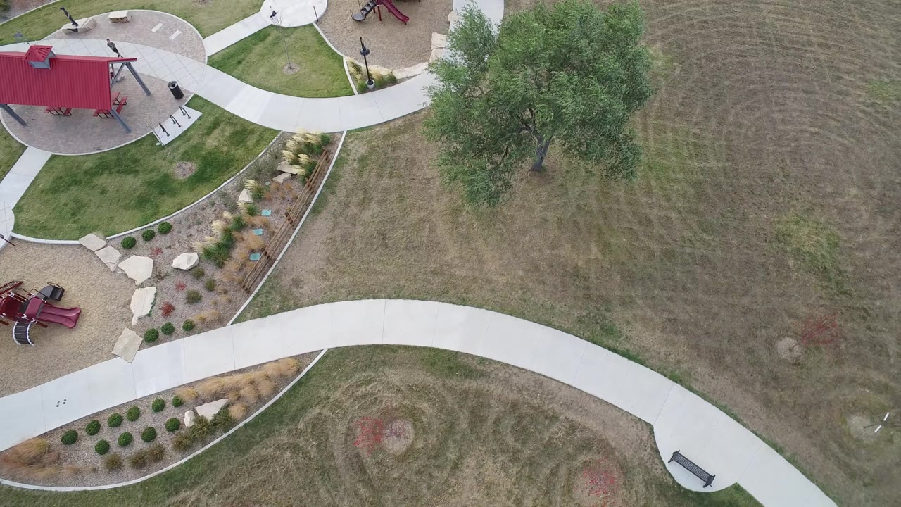 Overhead Hidden Lake, Colorado frame with the red structure near the upper left, pale walkways crossing through grass, planted beds below, a large tree on the right, and tiny people visible near the paths.