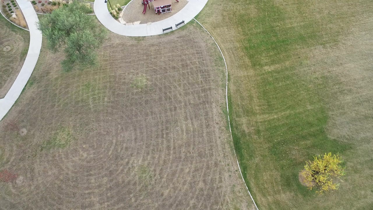 Overhead frame in Hidden Lake, Colorado showing a patchy lawn bordered by a curved concrete sidewalk, trees, benches, planting beds, and part of a red playground, with no visible people.