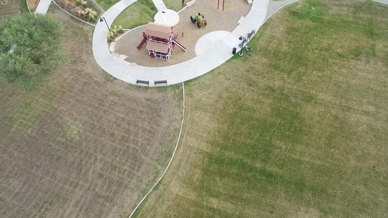 Overhead aerial frame in Hidden Lake, Colorado showing the top edge of a playground, curving path, benches, small trees, and a broad patchy grass field with no visible people.