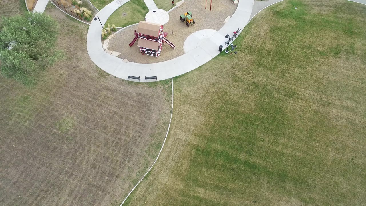 Hidden Lake, Colorado aerial view with the play structure near the top center, pale walkways wrapping the play area, and green-brown grass covering most of the frame with no visible people.