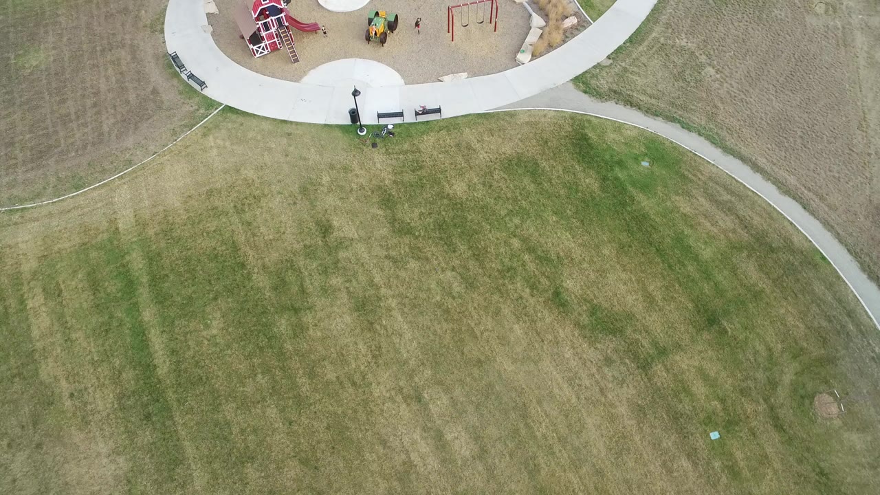Overhead frame from Hidden Lake, Colorado showing a mowed green and tan lawn below a curved concrete path and the edge of a playground, with no clearly visible people.
