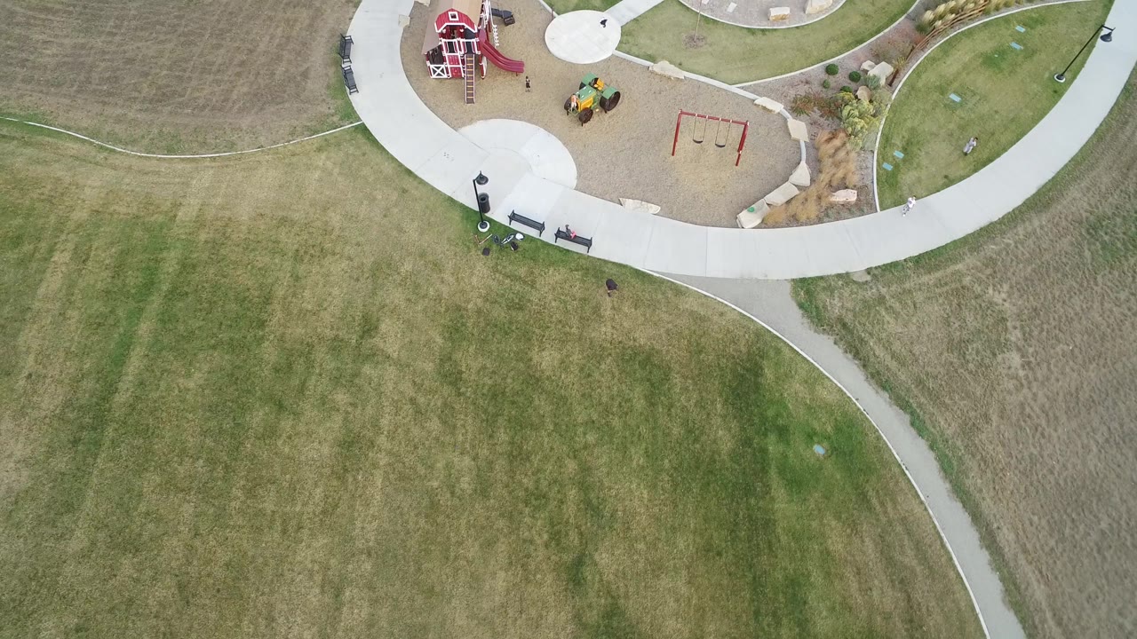 Top-down view over Hidden Lake, Colorado parkland with a red play structure near the top, red swings, pale concrete paths, open grass, benches, and small visible people.