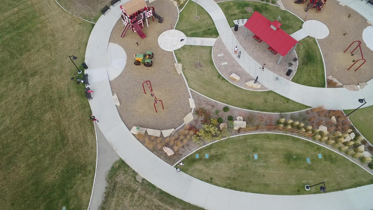 Overhead view of a park with curved paths, playground areas, lawns, and a red-roofed shelter.