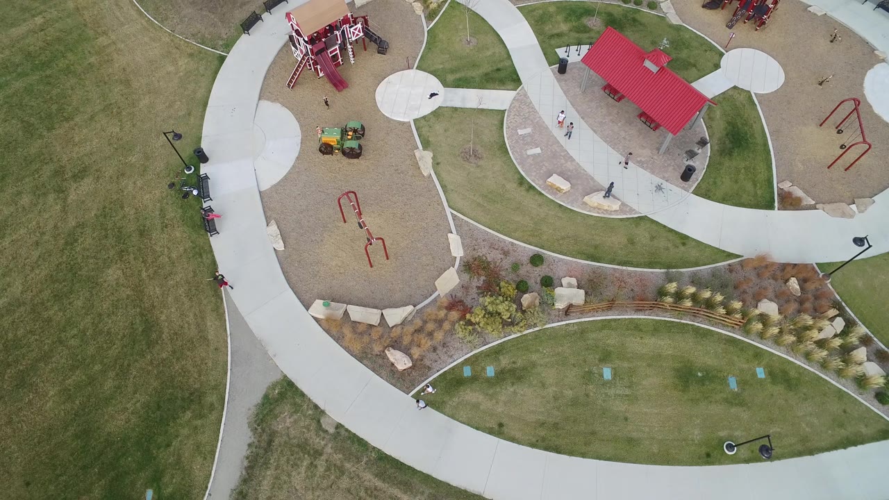 Second overhead frame in Hidden Lake, Colorado with the red shelter near the upper right, red play equipment on tan surfaces, broad lawns, curved paths, and tiny distant people along the walkways.