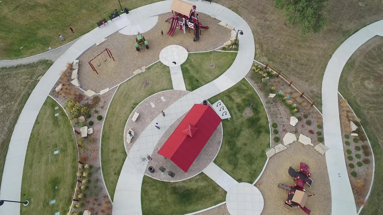Aerial frame of the Hidden Lake park with the red-roofed shelter slightly lower in view, pale paths looping around playground zones, landscaped beds, and small figures on the paved area.