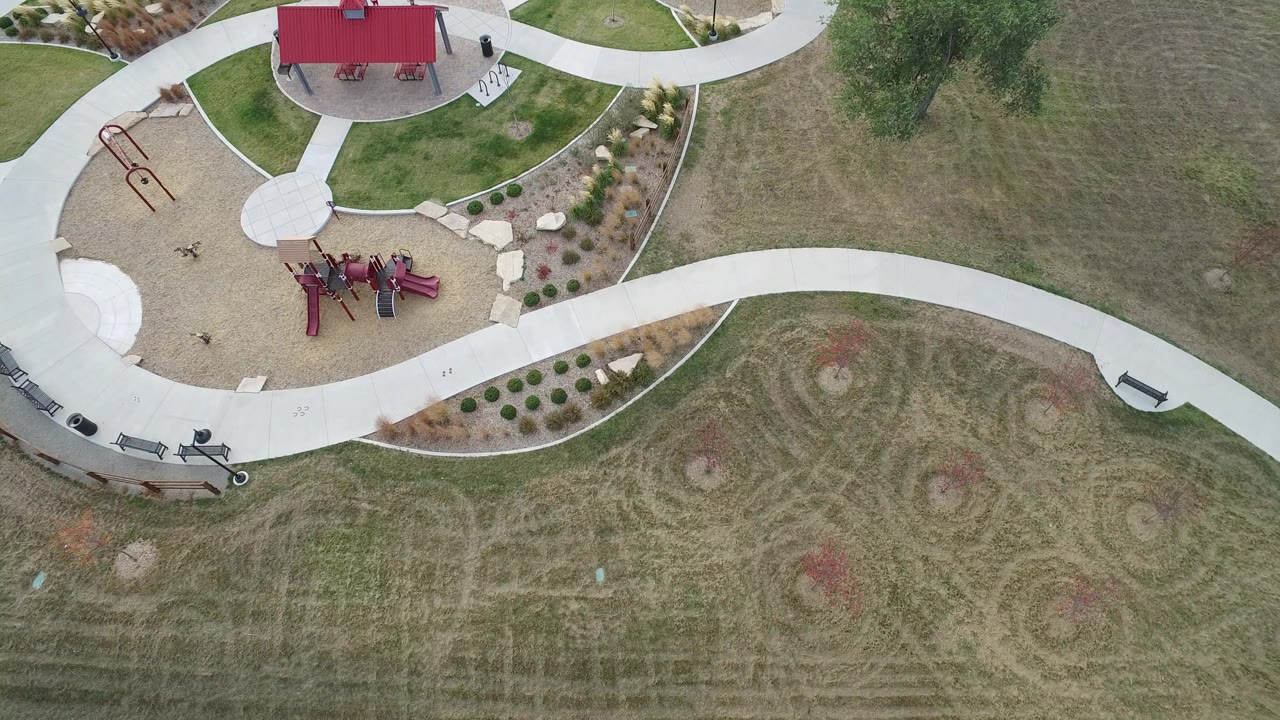 Top-down aerial frame from Hidden Lake, Colorado showing curved white paths, red playground equipment, mowed grass, benches, and a red-roof shelter with no visible people.
