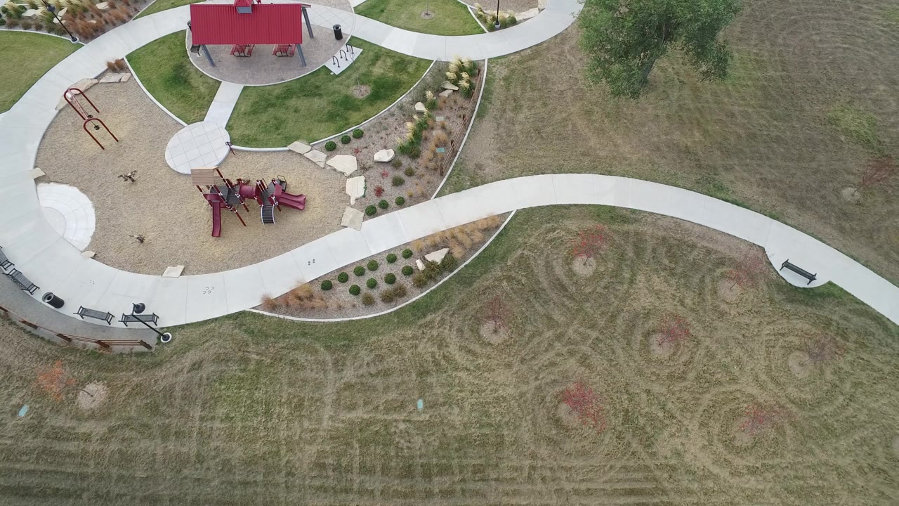 Overhead view of a park lawn with circular mowing marks, a playground, benches, and a red shelter roof.