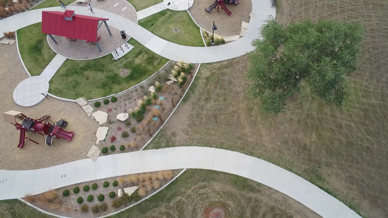 Overhead view of a small park with red playground structures, curved paths, grass, and a large tree.
