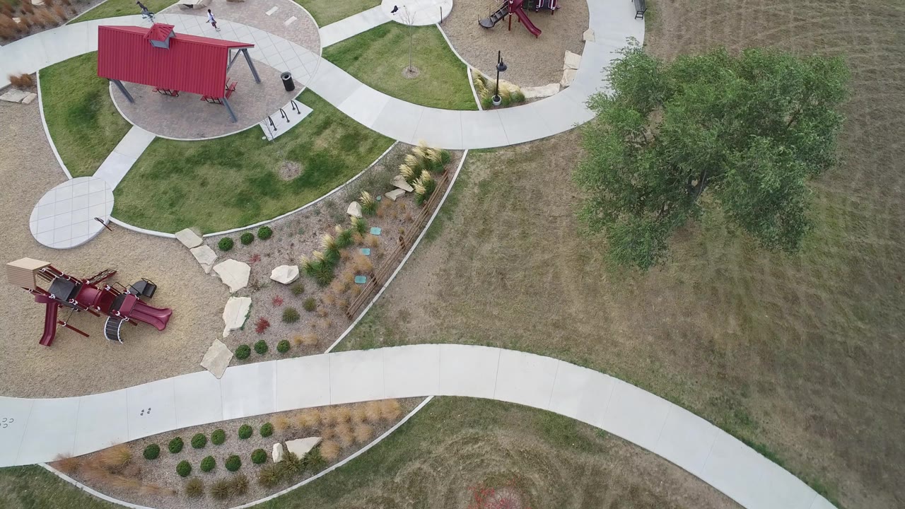 High aerial frame over the Hidden Lake, Colorado park, with the red roofed play structure near the top, a slide area at lower left, curved concrete paths, benches, and a large tree to the right.