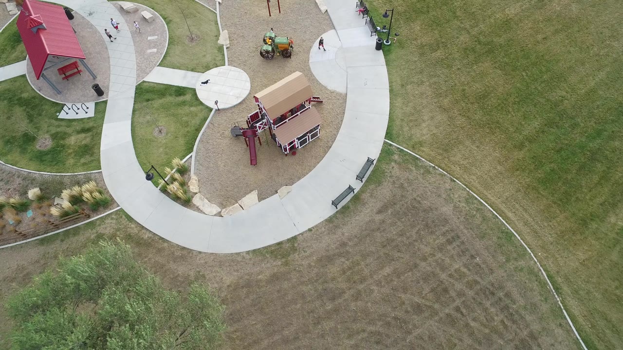 Aerial frame from Hidden Lake, Colorado with the red-roof shelter and playground near the top, pale concrete paths crossing the grass, benches, and small people visible.