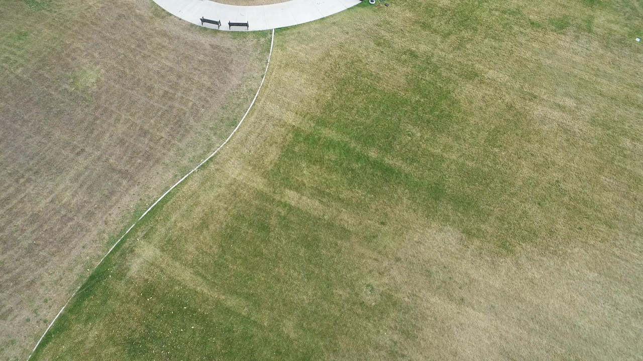 High aerial frame over a patchy green and tan grass field in Hidden Lake, Colorado, with a curved pale path and benches near the top edge, no visible people.