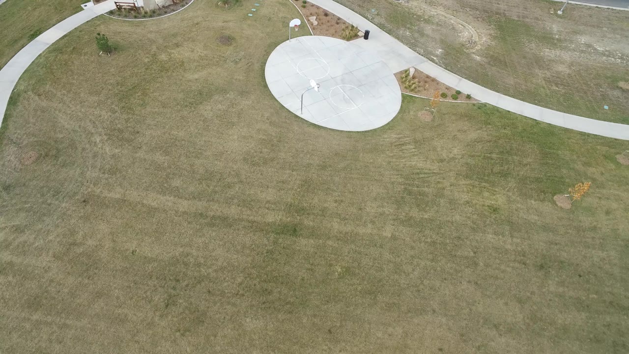 Aerial view in Hidden Lake, Colorado with open grass dominating the frame and a pale circular court, path, planting bed, and no visible people near the upper edge.