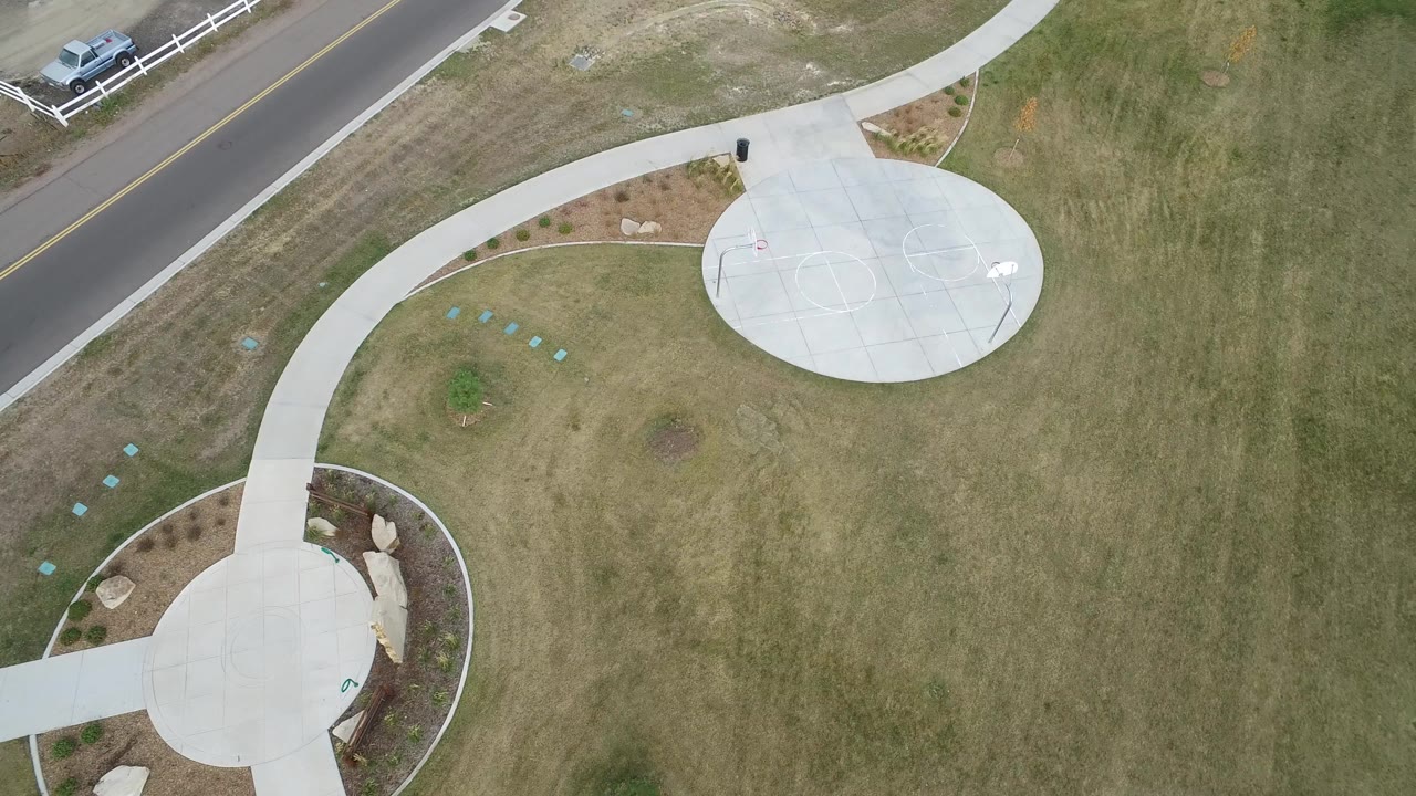 Overhead 2016 Hidden Lake, Colorado frame showing two round concrete pads connected by curved sidewalks beside grass, a road, parked vehicles, white fencing, and no visible people.