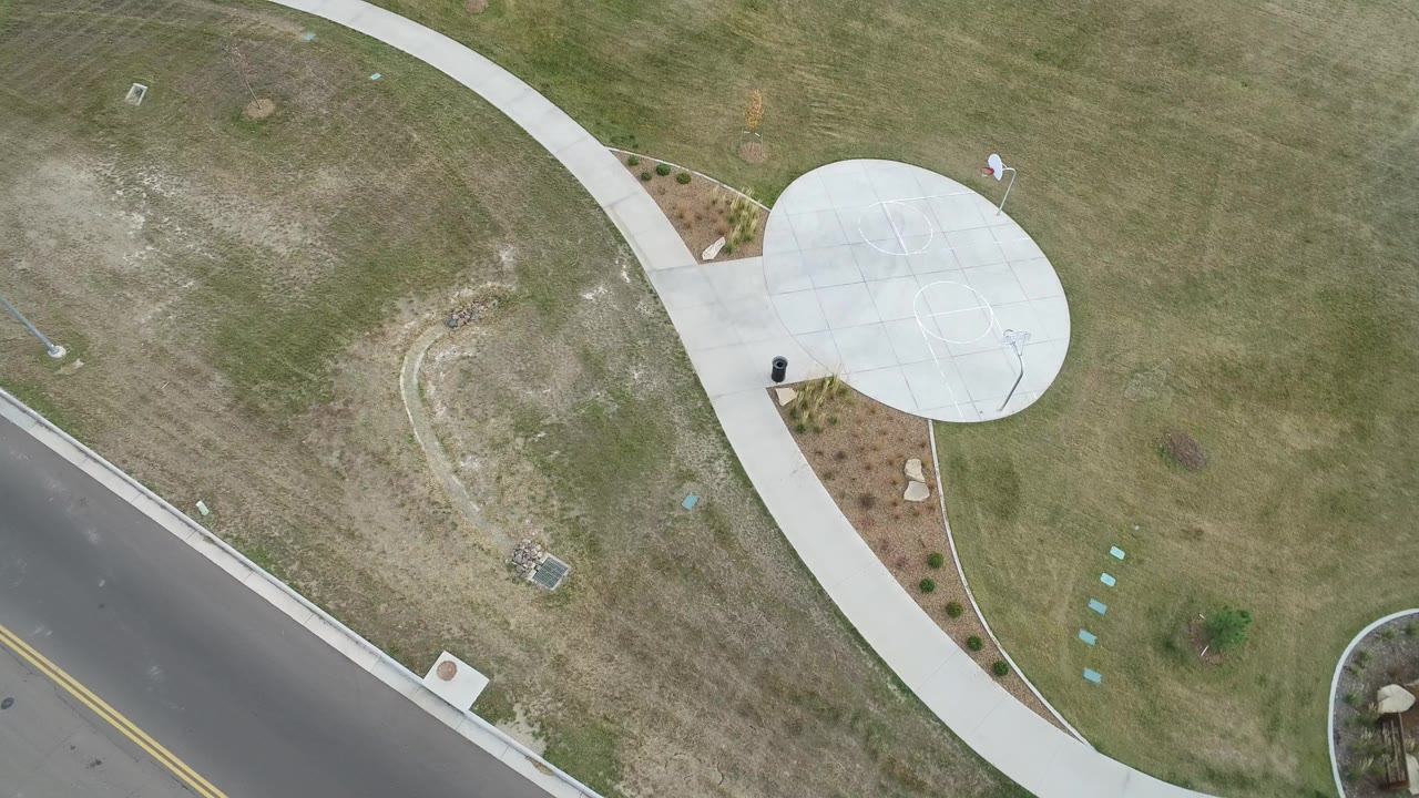 Overhead frame from Hidden Lake, Colorado showing a curved white concrete path, mowed grass, a two-lane road at lower left, and a circular paved area near the upper right.