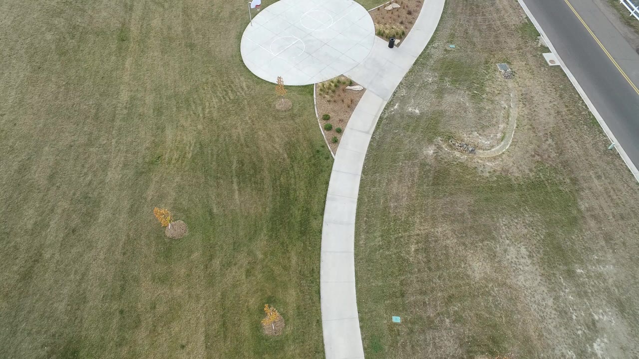 Overhead frame in Hidden Lake, Colorado showing mowed green and tan grass, a curved concrete sidewalk, a circular concrete pad, planted beds, and a paved road edge with no visible people.