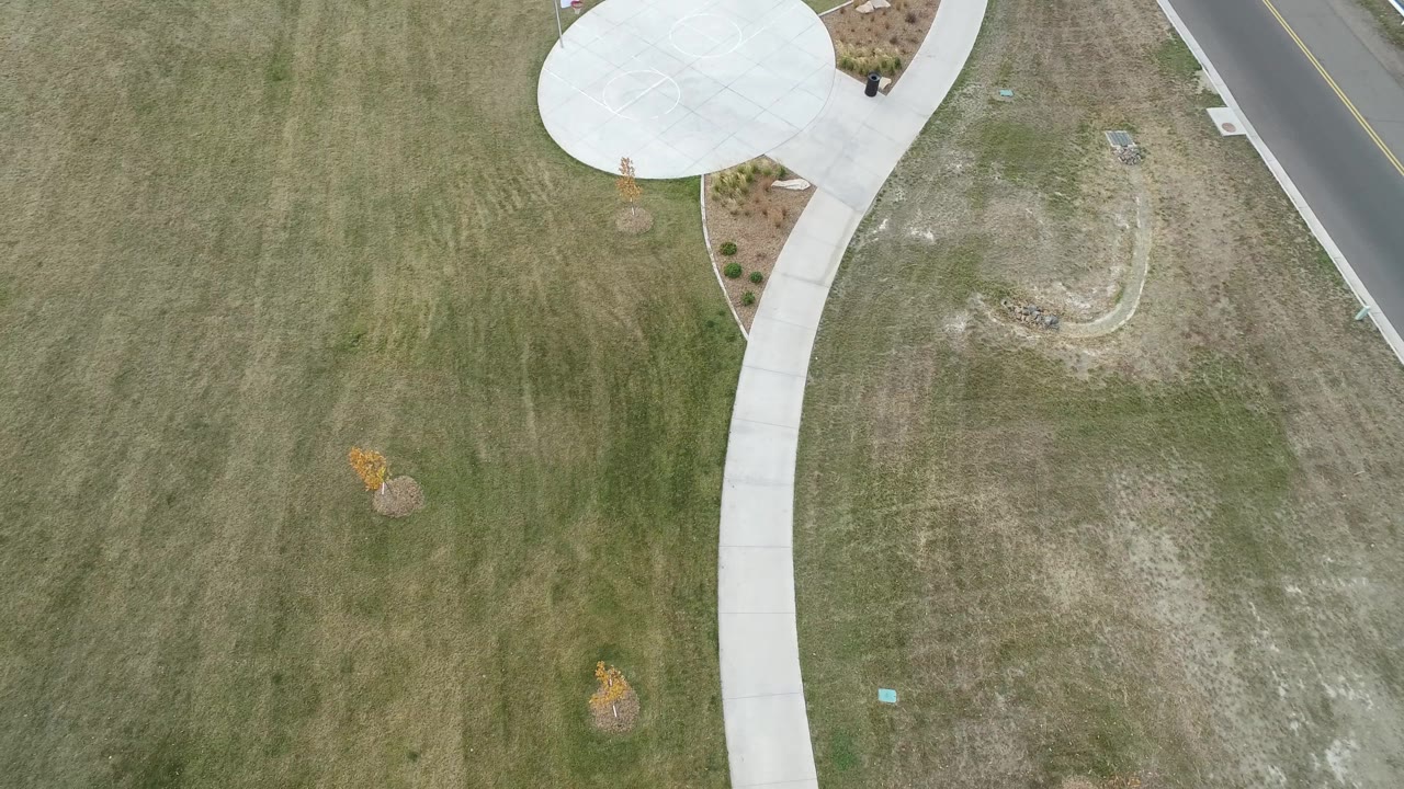 Top-down grass field with sidewalk, circular pad, landscaped beds, and roadside pavement.