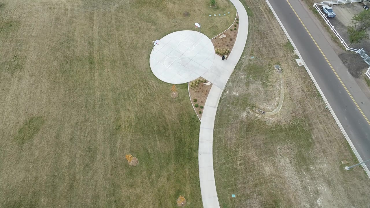 High overhead view in Hidden Lake, Colorado showing muted grass, a round concrete pad, a curving sidewalk, part of a two-lane road, and no visible people.