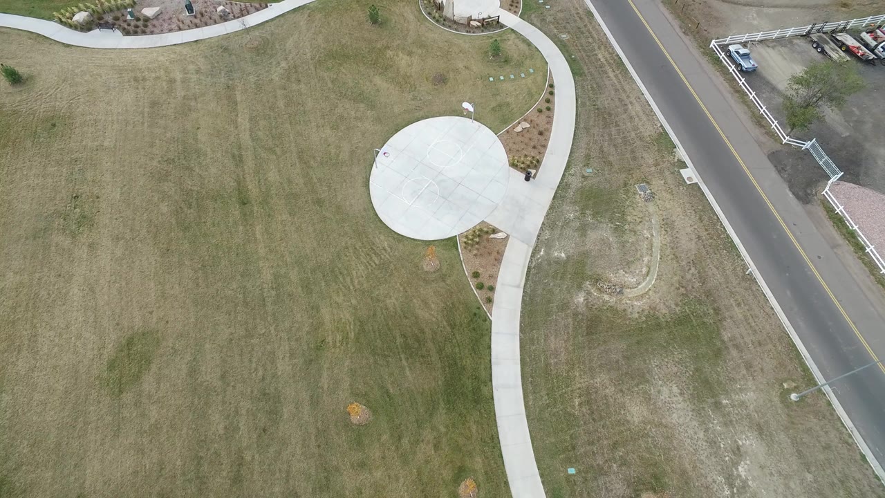 Aerial view of grass, a curving sidewalk, a round concrete pad, road, and parked cars.