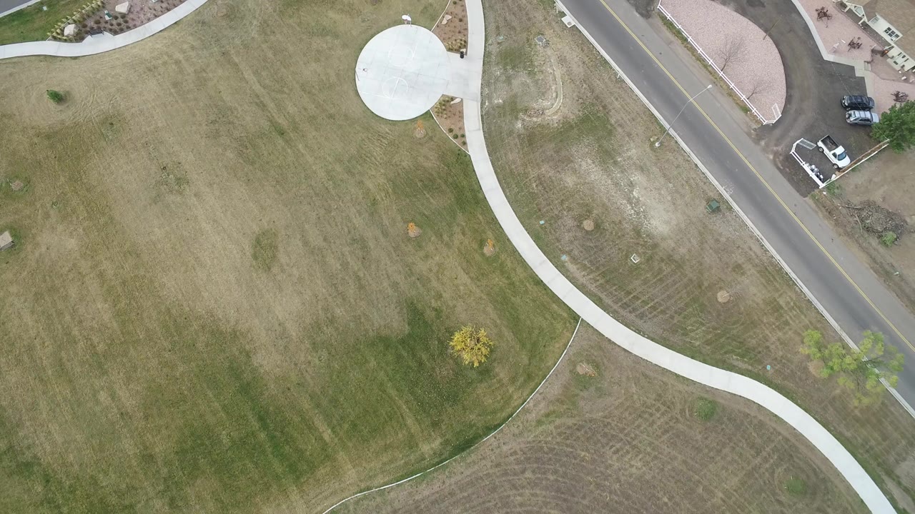 High overhead frame in Hidden Lake, Colorado showing tan and green grass, a curved white sidewalk, a road at the upper right, small trees, parked vehicles, and no visible people.