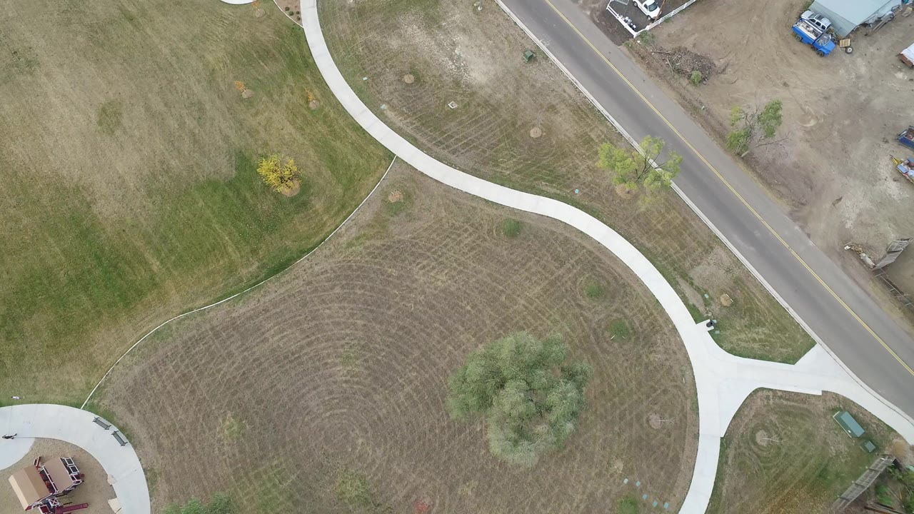 High aerial frame over Hidden Lake, Colorado showing a pale curving sidewalk through green-brown grass, a roadside edge, small yard structures, and no visible people.