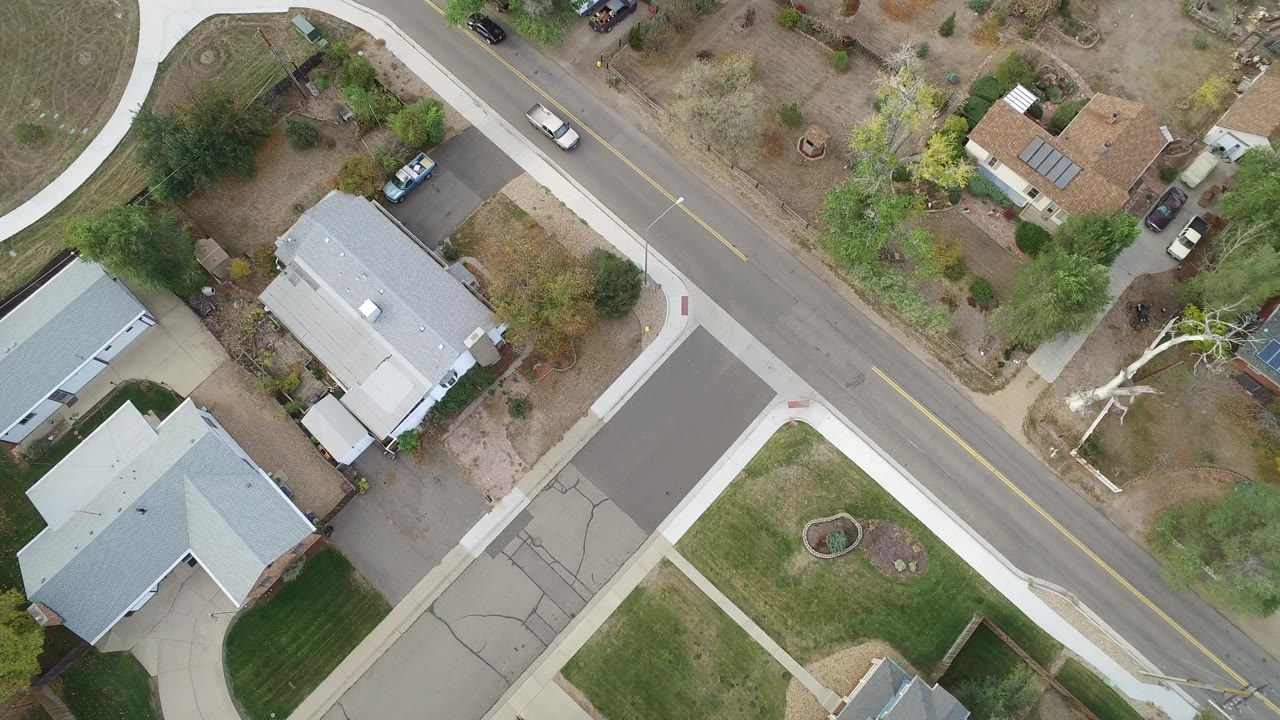 Aerial frame over Hidden Lake, Colorado showing the road cutting diagonally past homes, sidewalks, mixed dry and green yards, parked cars, and a light-colored car progressing along the lane, with no visible people.