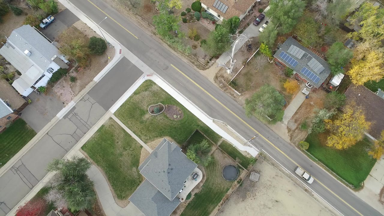 Overhead view of a residential street, houses, lawns, trees, and parked cars.