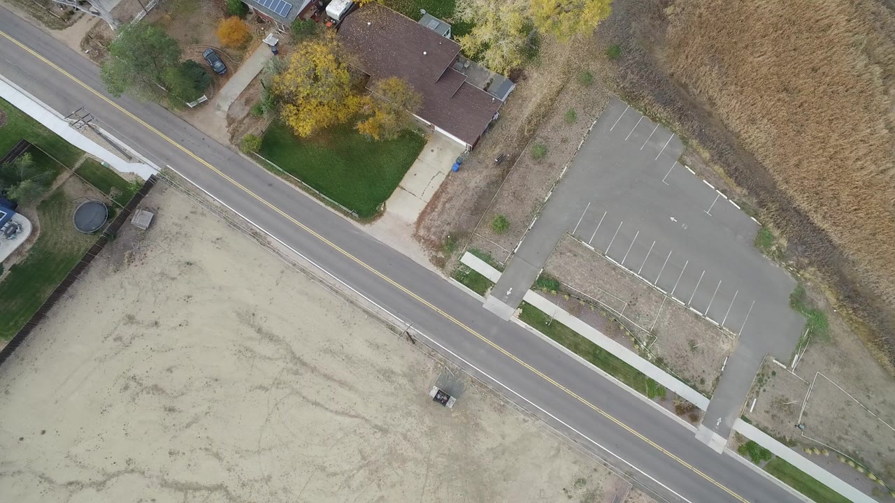 Top-down view of a road beside homes, dirt lots, grass, and an empty parking lot.