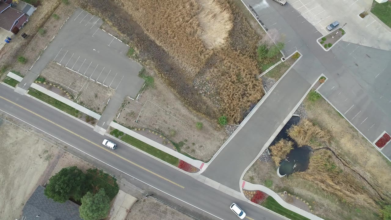 Top-down aerial view of a road, parking lot, pond, dry grass, and small vehicles.