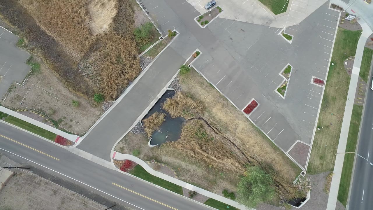 High overhead frame in Hidden Lake, Colorado showing a mostly empty parking lot, curved road edge, sidewalks, dry vegetation, a small pond, and no visible people.