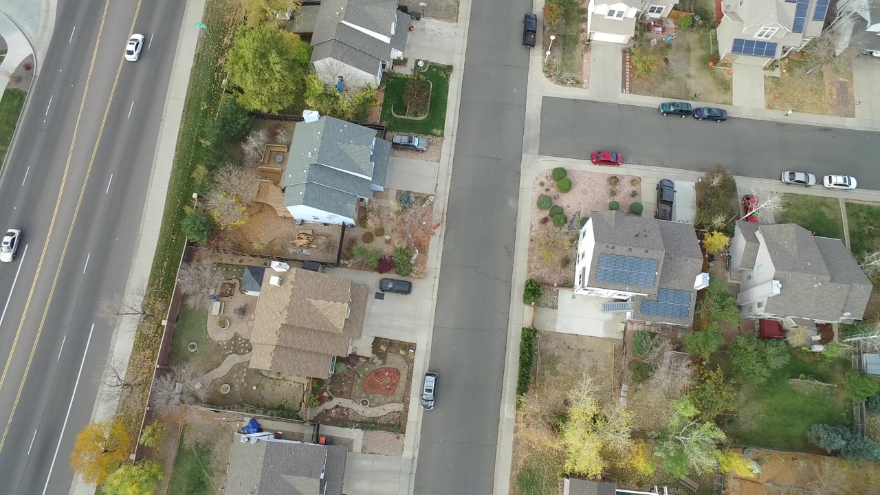 Overhead Hidden Lake, Colorado view centered on a residential block with detached houses, pale driveways, parked vehicles, lawns, bare trees, and a broader road along the left edge.