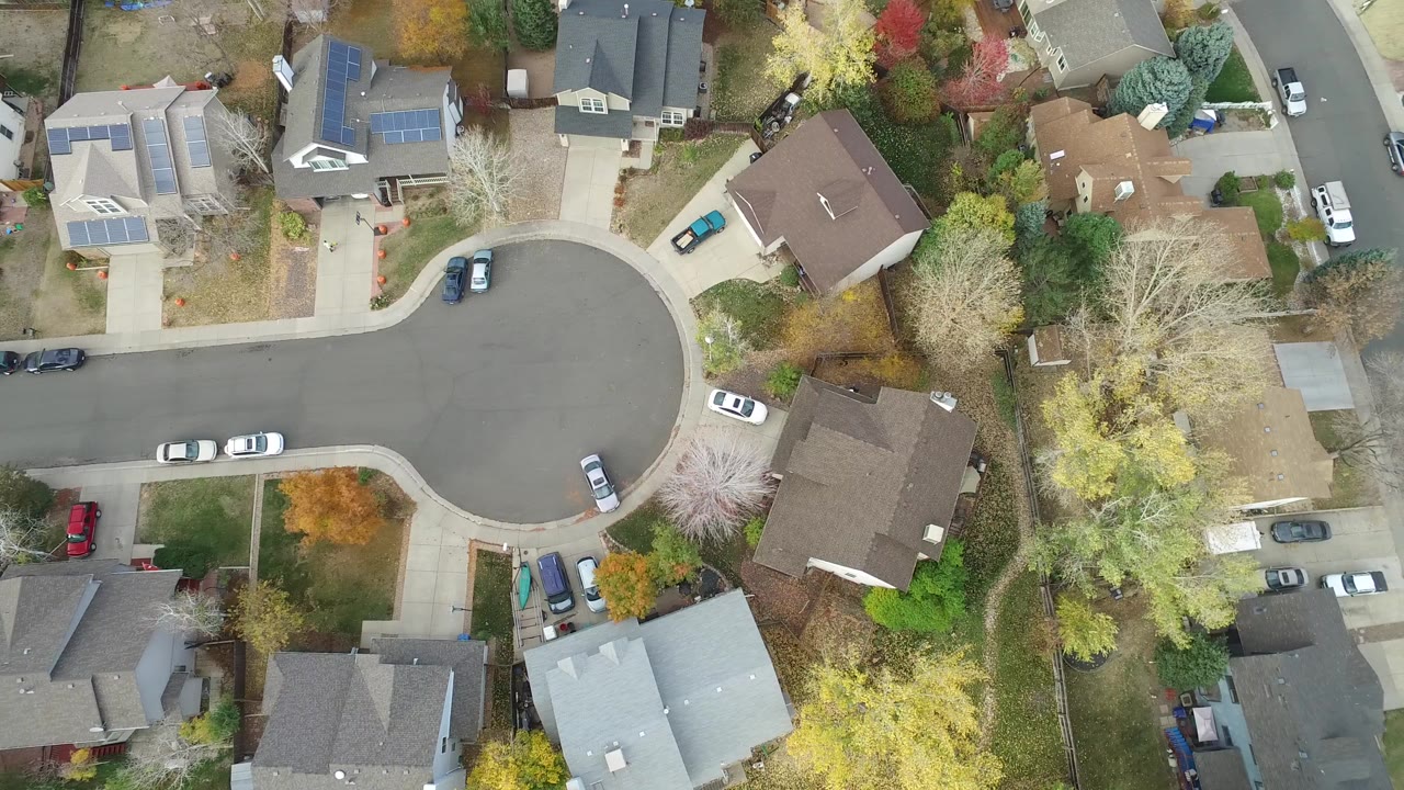 Overhead drone view in Hidden Lake, Colorado showing a residential cul-de-sac bordered by detached homes, driveways, parked cars, lawns, and no visible people.