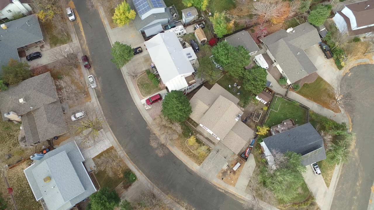Aerial frame over Hidden Lake, Colorado rooftops and yards, with a broad street curve on the left and parked vehicles near driveways, no visible people.