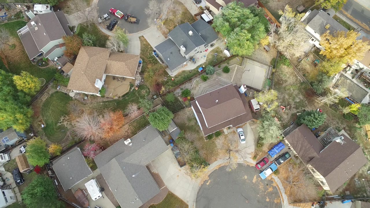 A high aerial view in Hidden Lake, Colorado shows a curving street edge, multiple rooflines, backyard fencing, parked vehicles, and no visible people.