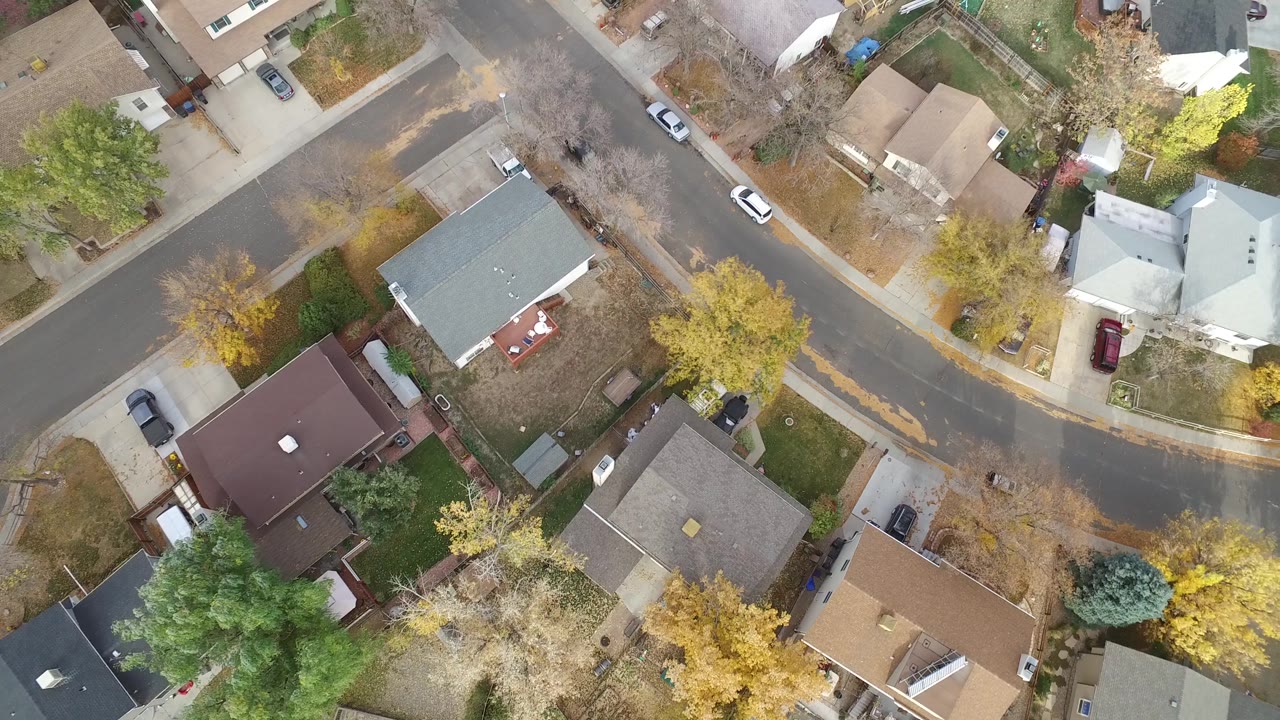 Overhead frame of Hidden Lake, Colorado residential lots with gray, brown, and light roofs around a curving road, parked vehicles, and yellow autumn trees, with no visible people.
