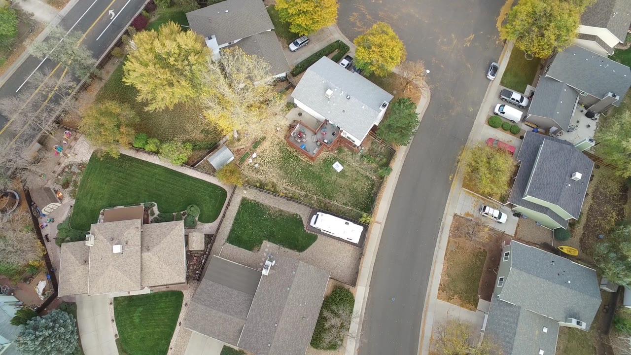 Aerial plan view in Hidden Lake, Colorado with a large tree near the center, houses around trimmed lawns, parked cars along driveways, and no visible people.