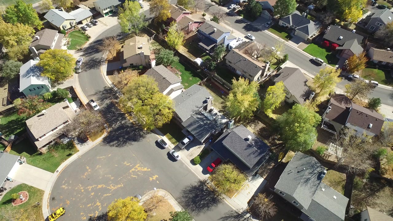 Drone-style overhead frame in Hidden Lake, Colorado showing a bend in the residential street, multiple rooftops, shaded yards, parked vehicles, and no visible people.