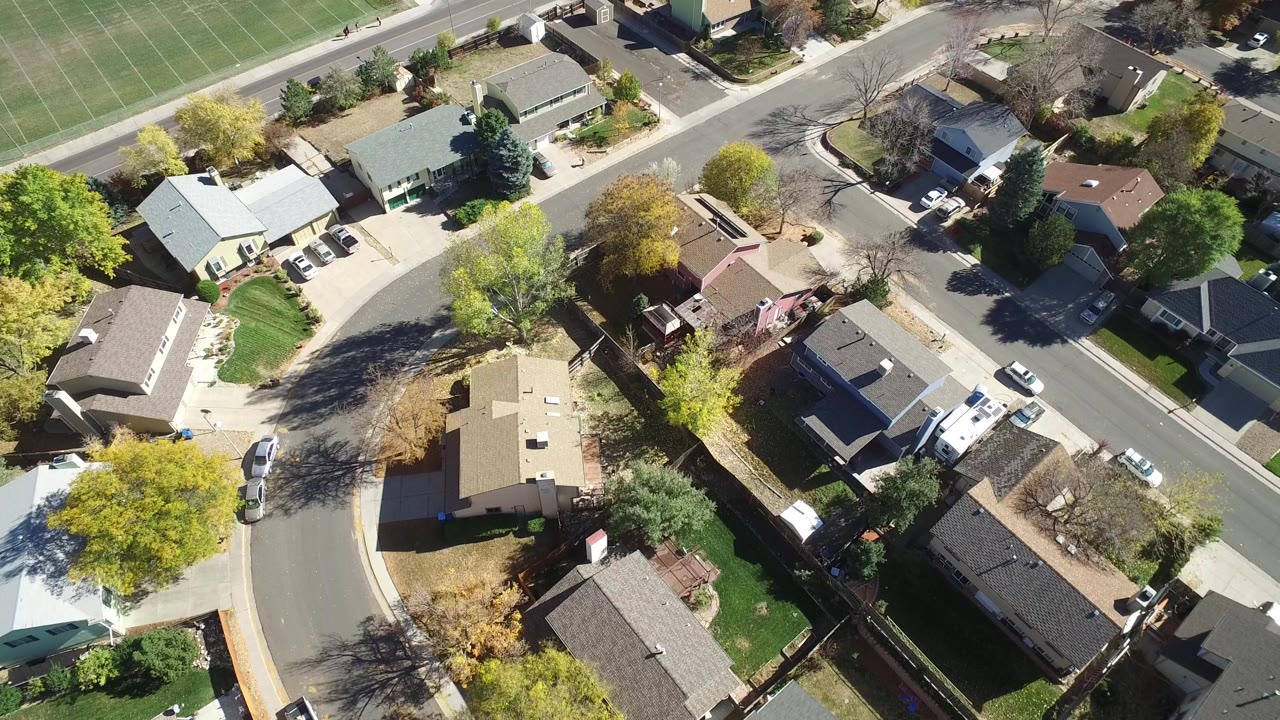 Aerial frame over Hidden Lake, Colorado with rooftops, a straight road, a green sports field at the upper edge, parked vehicles, and no visible people.