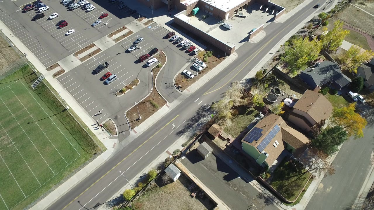 High aerial frame over Hidden Lake, Colorado showing a semi-empty parking lot, road lanes, nearby homes, trees, and the green edge of a sports field, with no clearly visible people.
